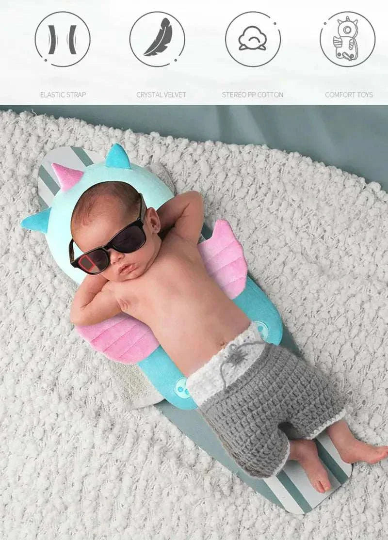 Baby wearing sunglasses lying on a soft blue and pink baby head protector pillow shaped like a dragon on a gray blanket